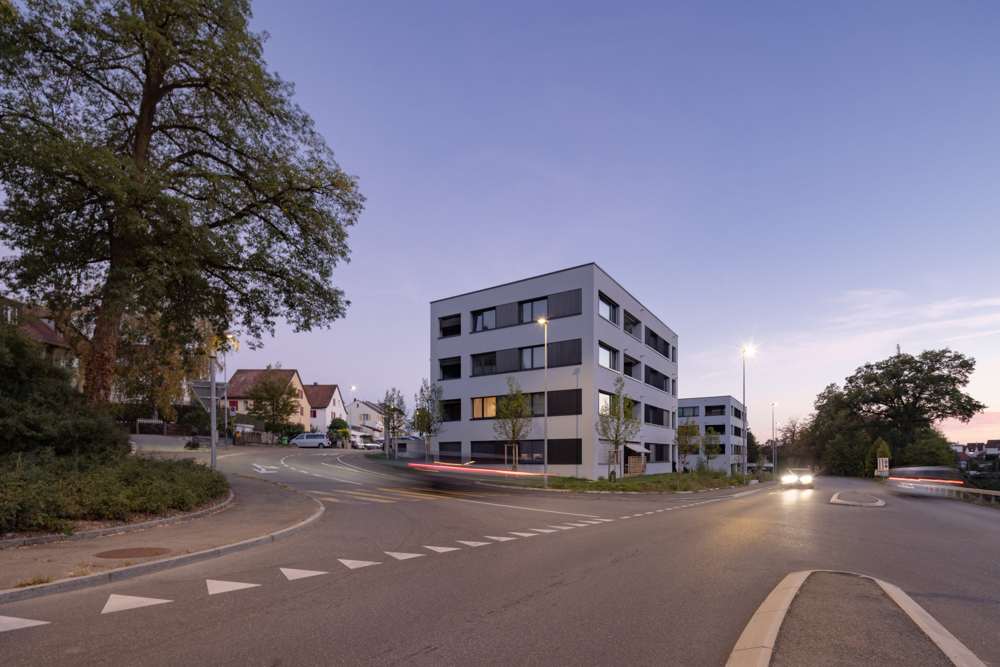 Die Anlage am Abend von der Strassenkreuzung: Die hellen Putzfassaden stehen ruhig im Strassenraum, die dunklen Paneele und Stoffbänder zeichnen die Geschosse nach.