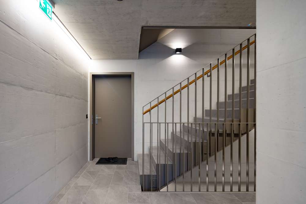 Eingangsgeschoss: Sichtbeton in Wänden und Decke, Boden und Stufen in Feinsteinzeug, Lichtband unter der Decke, Wohnungstüren im Ton der Geländer.
