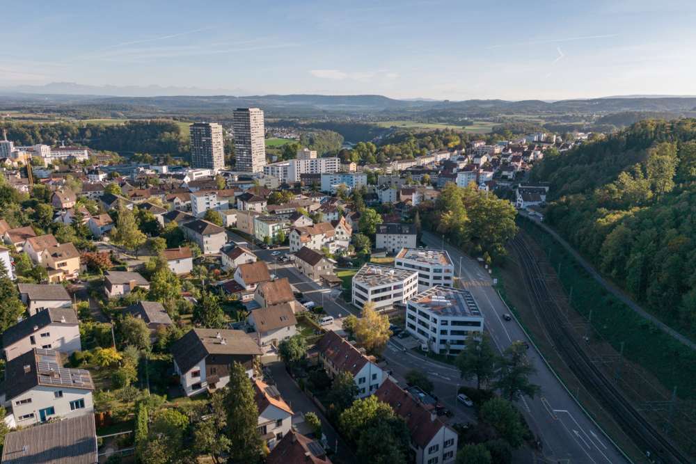 Luftaufnahme: Die drei Häuser mit ihren Flachdächern zwischen Strasse und Bahnlinie; im Hintergrund die Hochhäuser der Stadt.