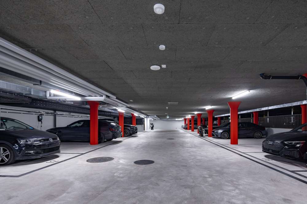 Einstellhalle im Überbaurecht: Stützen rot gestrichen, Holzwolleplatten an der Decke, Ladeinfrastruktur an der Wand.