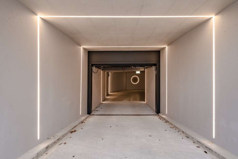 Einfahrt zur Tiefgarage: Lichtlinien zeichnen die Kanten des Sichtbetonraums nach; das Tor als dunkler Rahmen, dahinter die Ringleuchte der Garage.